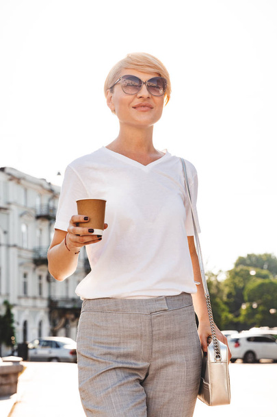 Photo of adult blond woman wearing white t-shirt and sunglasses walking through city street in summer and drinking takeaway coffee from paper cup - Zdjęcie, obraz