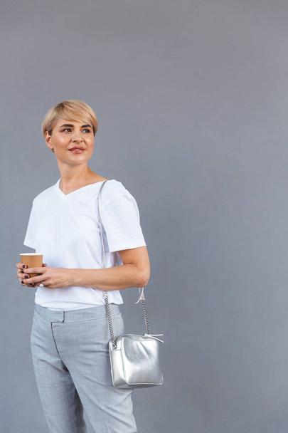Beautiful adult woman wearing white t-shirt standing isolated over gray wall and holding takeaway coffee - Fotoğraf, Görsel