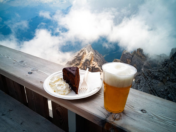 ブロンドのビールとザッハトルテ、山、岩、背景の雲のスライス - 写真・画像