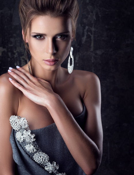 Portrait of young beautiful woman with sensual look. Professional makeup and large earrings. The hand on the shoulder. Evening dress with bare shoulders. The dark background - Zdjęcie, obraz