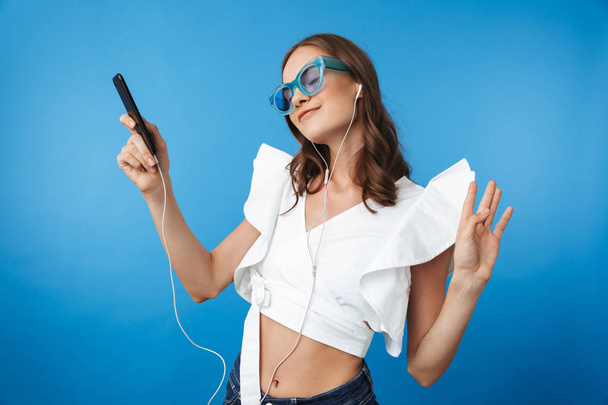 Portrait of a delighted young girl in summer clothes wearing sunglasses listening to music with earphones and mobile phone isolated over blue background - Φωτογραφία, εικόνα