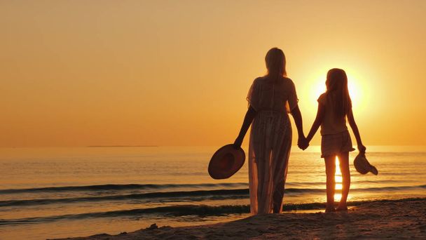 Μια γυναίκα κρατά ένα χέρι childs, περίπτερο μαζί στην παραλία, κοιτάξτε στο όμορφο ηλιοβασίλεμα. Μητέρα με την κόρη - Φωτογραφία, εικόνα