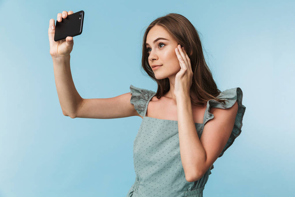 Portrait of a lovely young woman in dress taking a selfie isolated over blue background - Zdjęcie, obraz