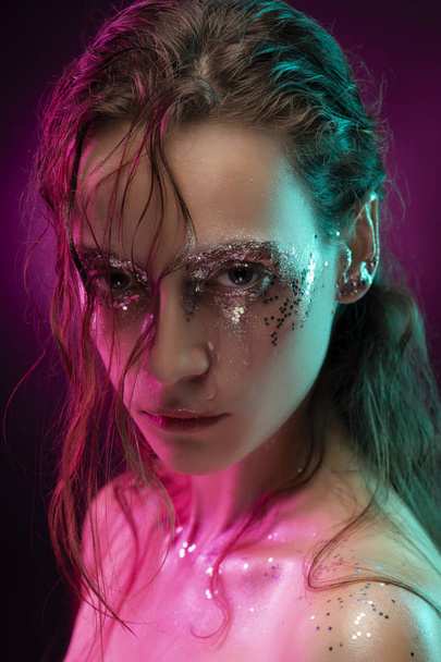Beautiful girl with creative makeup made of glitter with tears on her face illuminated with pink and blue light. Pink background. Conceptual, advertising and commercial design. Copy space. - Foto, afbeelding