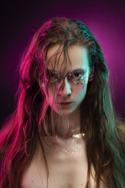 Beautiful girl with creative makeup made of glitter with tears on her face illuminated with pink and blue light. Pink background. Conceptual, advertising and commercial design. Copy space. - Φωτογραφία, εικόνα