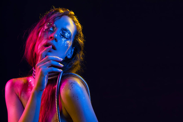 Beautiful girl with cretative make-up made of glitter with tears on her face wraps her neck with a metallic spiral hose illuminated with pink, blue and yellow light. Isolated on black. Copy space. - Foto, imagen