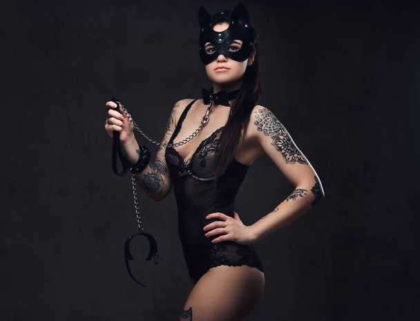 Σαγηνευτική αισθησιακή ερωμένη κοπέλα φορώντας μαύρα εσώρουχα σε Bdsm εξαρτήματα και γάτα δερμάτινη μάσκα θέτοντας σε σκούρο φόντο. Απομονωμένα σε σκούρο φόντο.    - Φωτογραφία, εικόνα