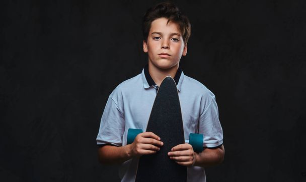 Portret van een jonge skater jongen gekleed in een wit t-shirt houdt een longboard. Geïsoleerd op een donkere achtergrond. - Foto, afbeelding
