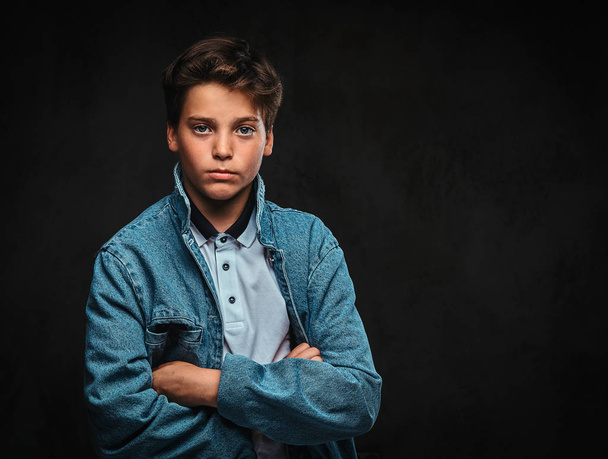 Fashionable young guy dressed in a t-shirt and denim jacket posing with crossed arms in a studio. Isolated on the dark background. - Photo, Image