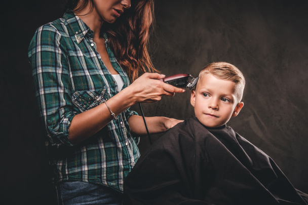 子供美容師トリマーには、暗い背景に小さな男の子を切っています。かわいい幼児少年取得散髪.  - 写真・画像