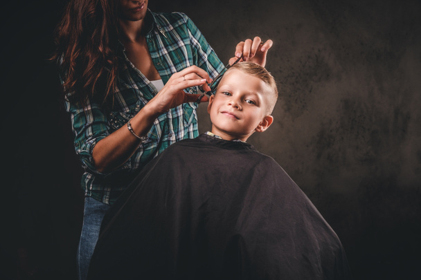 Çocuk Kuaför makası ile karanlık bir arka plan karşı küçük çocuk kesiyor. Saç kesimi elde memnun sevimli okul öncesi çocuk.  - Fotoğraf, Görsel