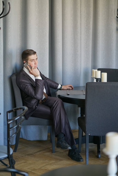 um jovem em um terno de negócios preto, camisa branca e gravata calmamente falando em um telefone celular, sentado em um café em uma mesa
 - Foto, Imagem
