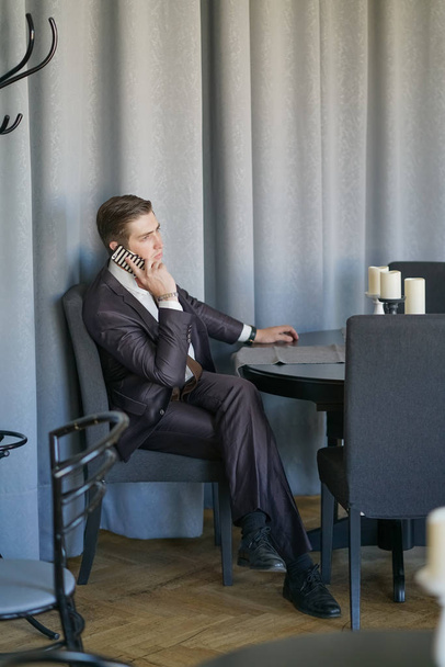 un joven en un traje de negocios negro, camisa blanca y corbata hablando tranquilamente en un teléfono celular, sentado en un café en una mesa
 - Foto, imagen