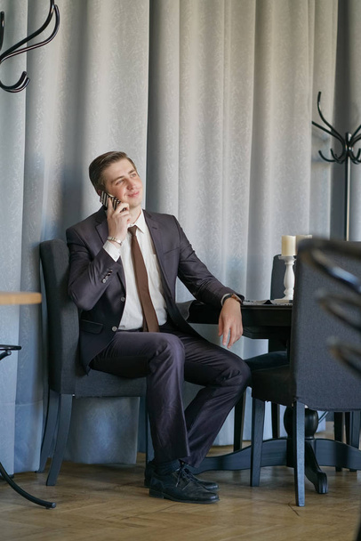 un joven en un traje de negocios negro, camisa blanca y corbata hablando tranquilamente en un teléfono celular, sentado en un café en una mesa
 - Foto, imagen