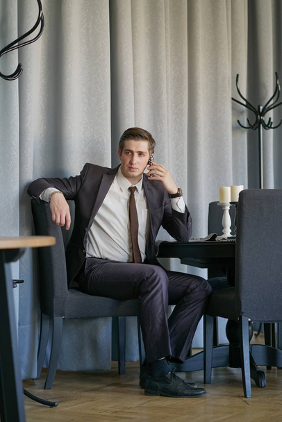 un giovane uomo in abito da lavoro nero, camicia bianca e cravatta parlare tranquillamente su un telefono cellulare, seduto in un caffè a un tavolo
 - Foto, immagini