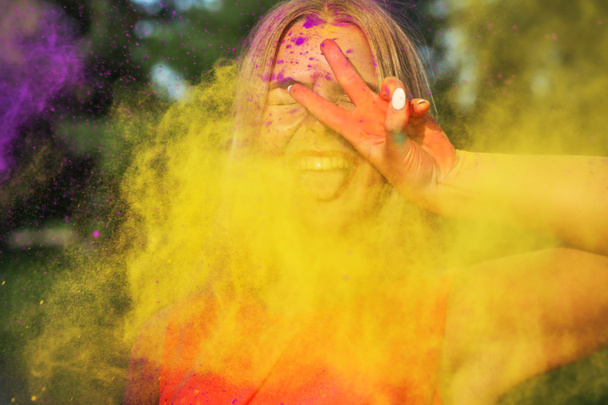 Laughing young woman standing in a cloud of yellow paint Holi showing peace gesture - Photo, Image