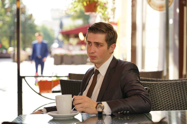 um jovem em um terno de negócios preto, camisa branca e gravata está sentado em um café de rua da cidade em uma mesa e desfrutar de uma xícara de café
 - Foto, Imagem