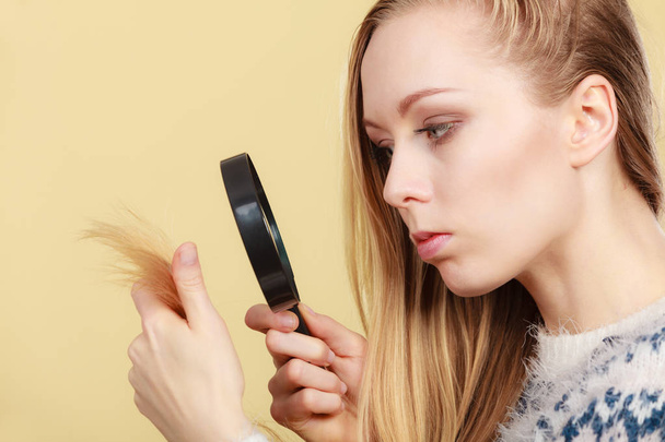 Haircare, health problem concept. Unhappy blonde woman looking at ends of her blonde hair through magnifying glass - Photo, Image