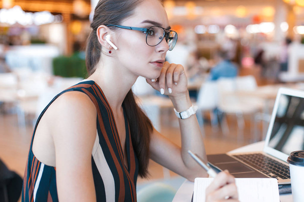 Aantrekkelijk meisje in glazen schrijft in notitieblok in café. Lifestyle business portret - Foto, afbeelding