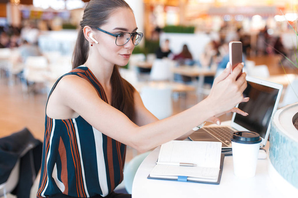 Молодая привлекательная девушка в очках делает селфи, используя телефон в кафе. Портрет в стиле жизни
 - Фото, изображение