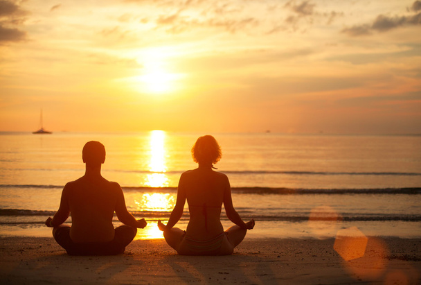 Um jovem casal sentado na praia do mar na posição de lótus ao pôr-do-sol. Prática de ioga
. - Foto, Imagem