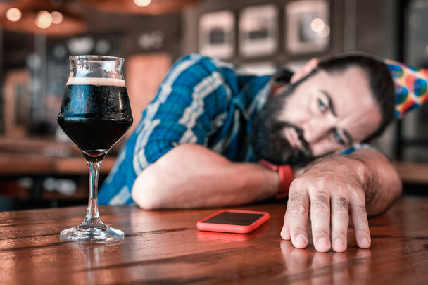 Bearded dark-haired man looking at glass of nice cold dark beer - Фото, изображение