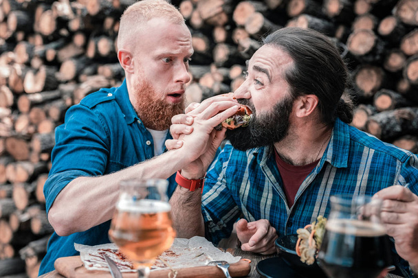 Hungry dark-haired man trying meat burger of his friend - Photo, image