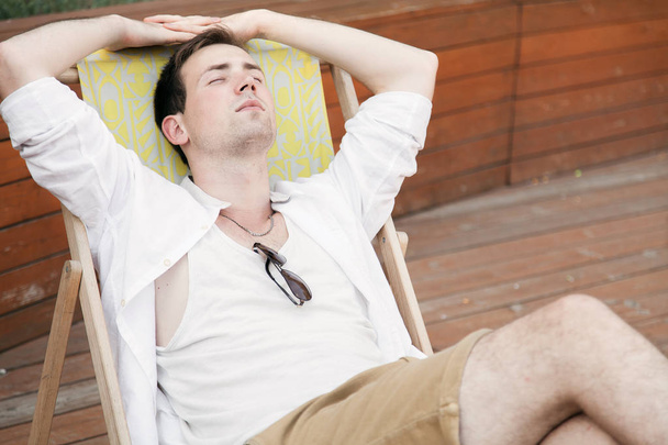 Ένας νεαρός άντρας κάθεται επάνω σε μια ξαπλώστρα στον καθαρό αέρα. Πορτρέτο. - Φωτογραφία, εικόνα