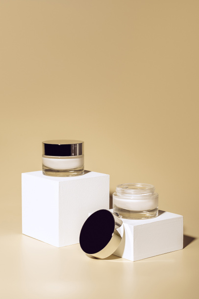 Vista de cerca de las cremas faciales y corporales en frascos de vidrio sobre cubos blancos sobre fondo beige
 - Foto, Imagen