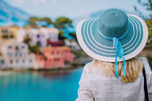 Rear view of tourist woman wear blue sunhat and white clothes admire view of colorful tranquil colorful houses of Assos village on sunny day. Visiting Kefalonia in summer. Travel vacation in Greece - Foto, afbeelding