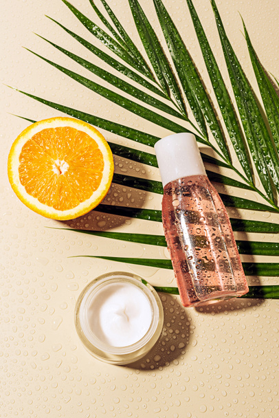 top view of facial cream and lotion, palm leaf and orange half with water drops on beige background - Valokuva, kuva