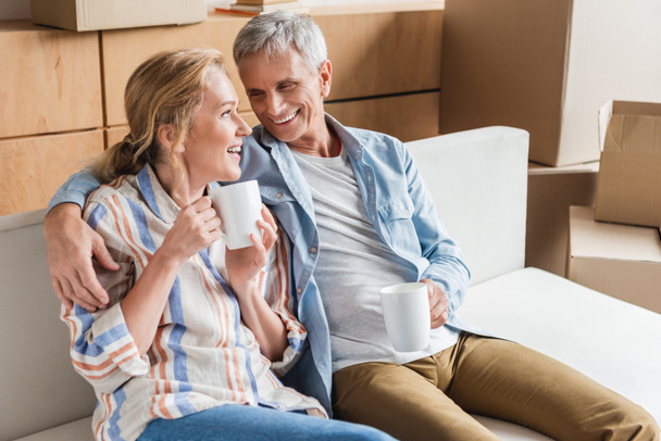 happy senior couple drinking tea and smiling each other while sitting on couch during relocation - Photo, Image