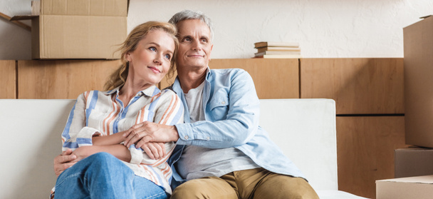 glückliches älteres Paar umarmt und schaut weg, während es zusammen auf der Couch im neuen Haus sitzt   - Foto, Bild