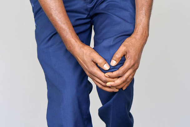 Douleur au genou. Gros plan Jambe masculine africaine avec genoux douloureux sur fond gris. Homme sentant la douleur articulaire, ayant des problèmes de santé et touchant jambe avec les mains. Concept de corps et de soins de santé
. - Photo, image