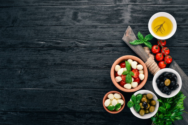 Ingredients for Italian caprese salad. Mozzarella cheese, cherry tomatoes, basil leaves, olives, oil, pepper. On a black wooden background. Free space for text. - Foto, immagini