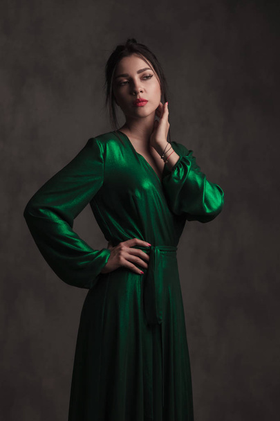 ritratto di giovane donna bruna che indossa un abito lungo verde in piedi su sfondo grigio con una mano sul fianco e guardando a lato
 - Foto, immagini
