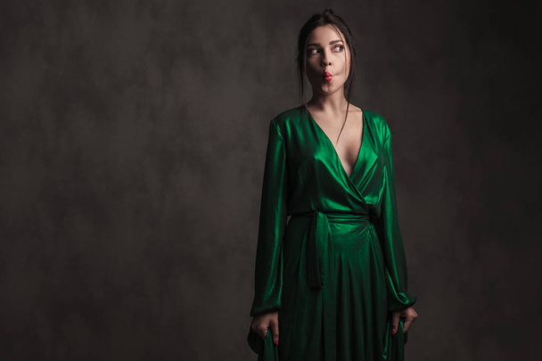 portrait of funny elegant woman wearing a long green dress making duck face and looking to side while standing on grey background - Photo, Image
