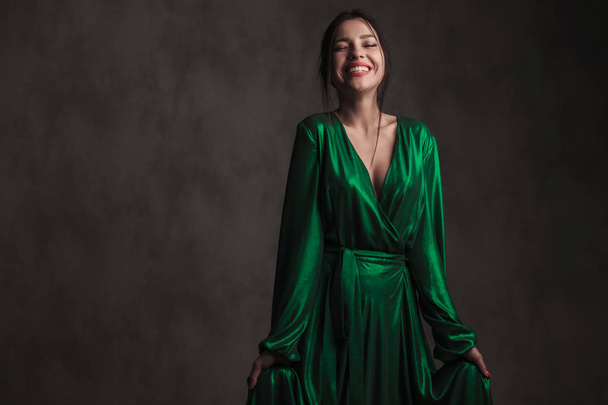 retrato de una hermosa mujer vestida con un vestido verde riendo mientras está de pie sobre fondo de walpaper gris, sosteniendo su vestido
 - Foto, imagen