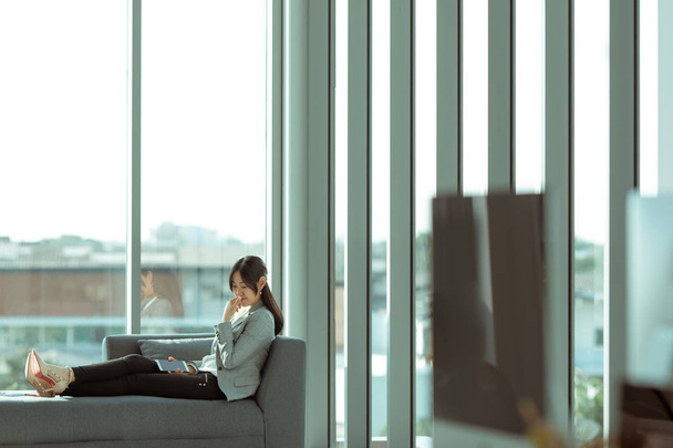 Une femme asiatique aux cheveux longs est assise sur le canapé se sentir seule dans une chambre moderne avec de grandes fenêtres à l'arrière
. - Photo, image