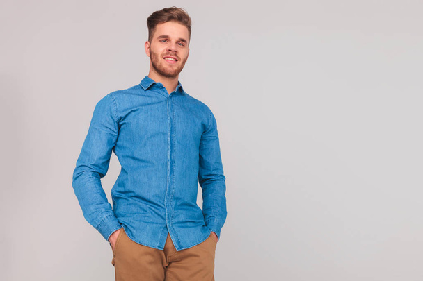 portrait of relaxed casual man in blue shirt standing on light grey background with hands in pockets - Photo, Image