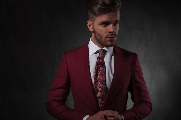 portrait of young man in red suit holding palms together while looking to side and standing on grey wallpaper background - Photo, Image