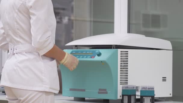 No laboratório, uma mulher de roupão de banho e luvas coloca amostras de células humanas em tubos de ensaio numa centrífuga para desenrolamento e separação do excesso de camadas de células
 - Filmagem, Vídeo