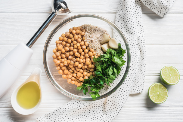 Flat lay with chickpeas, parsley and spices ingredients for hummus in bowl, blender and lime pieces on wooden surface - Foto, Imagen