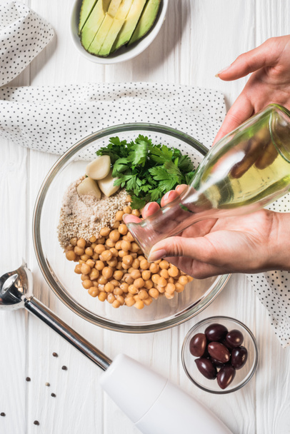 Foto recortada de mujer vertiendo aceite de oliva en un tazón con garbanzos y otros ingredientes para hummus en la mesa de madera
 - Foto, Imagen