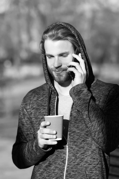 New technology and modern life. Guy with smartphone and takeaway drink on fresh air. Bearded man in hood hold disposable coffee cup on sunny outdoor. Coffee or tea mood. Morning tea for breakfast - Fotoğraf, Görsel