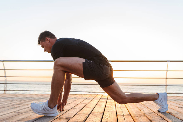 Energetic young sportsman doing stretching leg exercises at the seaside - 写真・画像