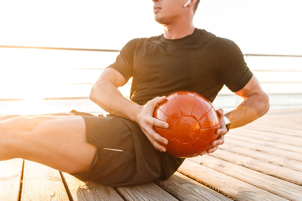 Cropped image of a young man in earphones doing exercises with a heavy ball outdoors - Foto, Bild