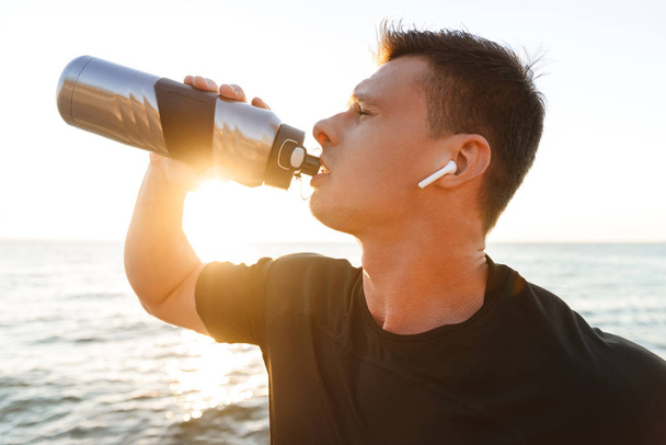 Exhausted young sportsman in earphones drinking water from a bottle at the seaside - 写真・画像