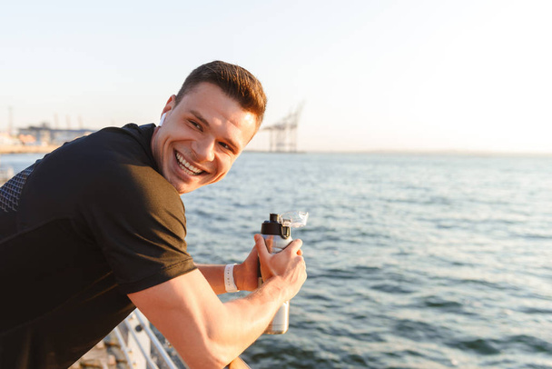 Happy young sportsman in earphones holding water bottle while leaning on rails at the seaside - Fotoğraf, Görsel