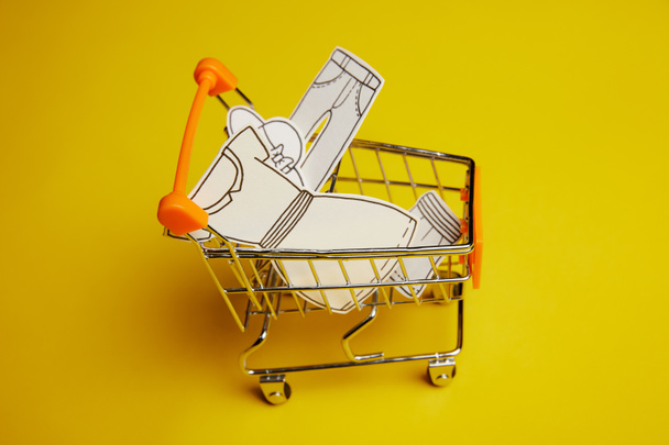 close up view of little shopping trolley with paper clothes on yellow backdrop - Fotografie, Obrázek